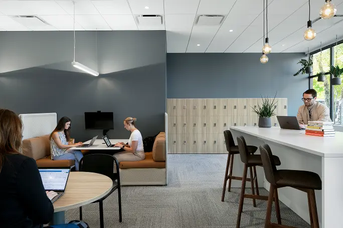 A modern office space featuring several workstations. In the foreground, a woman is seated at a round table using a laptop. Two women are working on laptops in a cozy seating area on the left. On the right, a man is seated at a counter with a laptop and books nearby. The background includes lockers and plants, with overhead lighting creating a bright atmosphere.