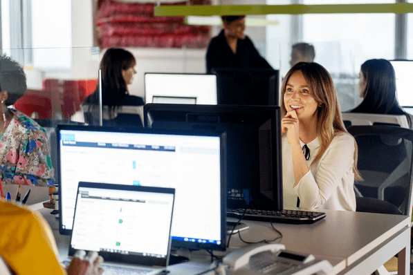A modern office environment with multiple people working at their desks. In the foreground, a woman with long hair is smiling, resting her chin on her hand while looking at the camera. There are several computer monitors displaying data and various office supplies on the desks. Other colleagues are seated at their computers, and someone is standing in the background, engaged in a discussion. The space is bright and features large windows.