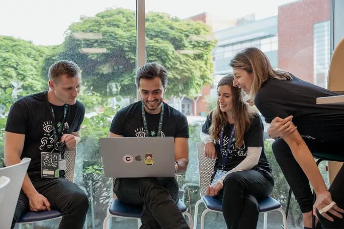 A group of four colleagues is gathered around a laptop, engaged in a discussion and reviewing content displayed on the screen. They are seated in a bright, open space with greenery visible through the large windows in the background. Each person is wearing a black t-shirt, and their expressions convey collaboration and enthusiasm as they cooperate on a project.