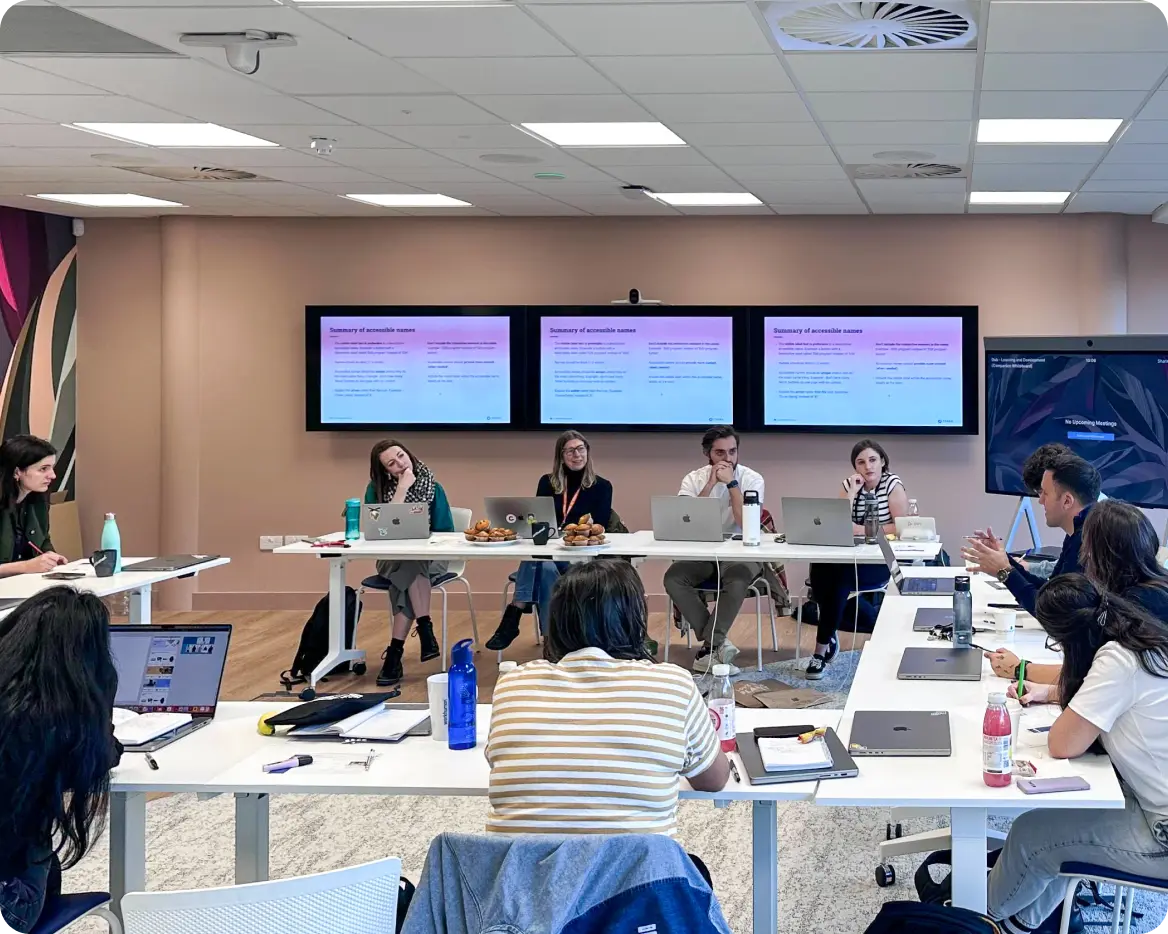 Accessibility annotation workshop in large meeting room: 4 employees lead the session with 7 attendees visible. 3 screens on rear wall show a presentation slide as the group turns to face a speaker.