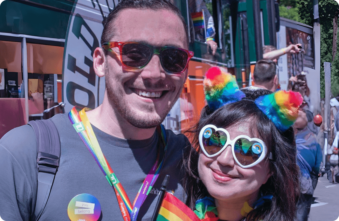 A smiling man and woman pose together at a lively event, both wearing colorful sunglasses and rainbow-themed accessories. The man has a gray shirt and a name badge, while the woman sports heart-shaped glasses and colorful ear accessories. They are standing outdoors, surrounded by a festive atmosphere, with people and vibrant decorations visible in the background. A button on the man's shirt reads "You Belong."