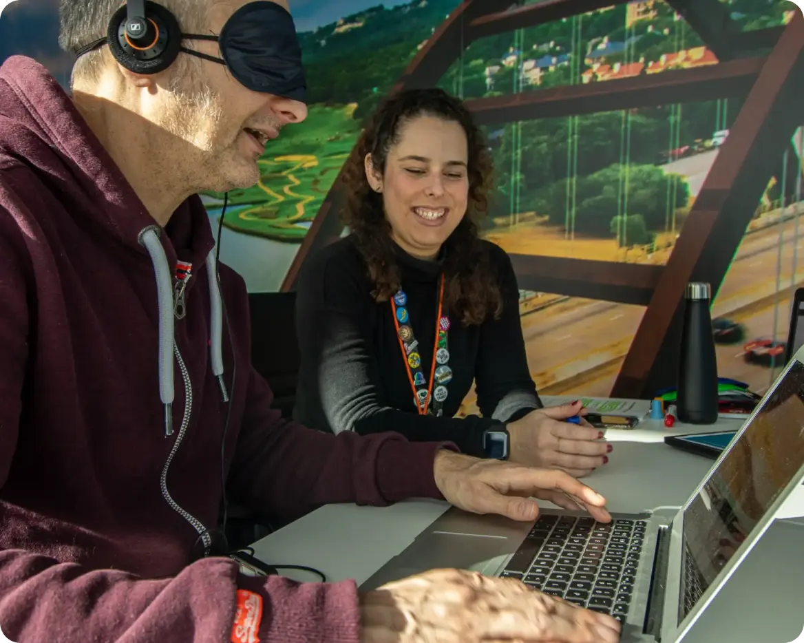 A Workhuman employee sits at a laptop wearing a blindfold and headphones. A smiling colleague sits with him and guides him through a screen reader activity for Usability day
