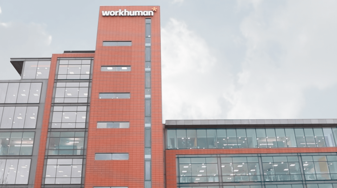 The image shows a building with a prominent sign for Workhuman. The structure features a modern design, with large glass windows and a combination of brick and glass exteriors. The sky in the background is slightly cloudy.