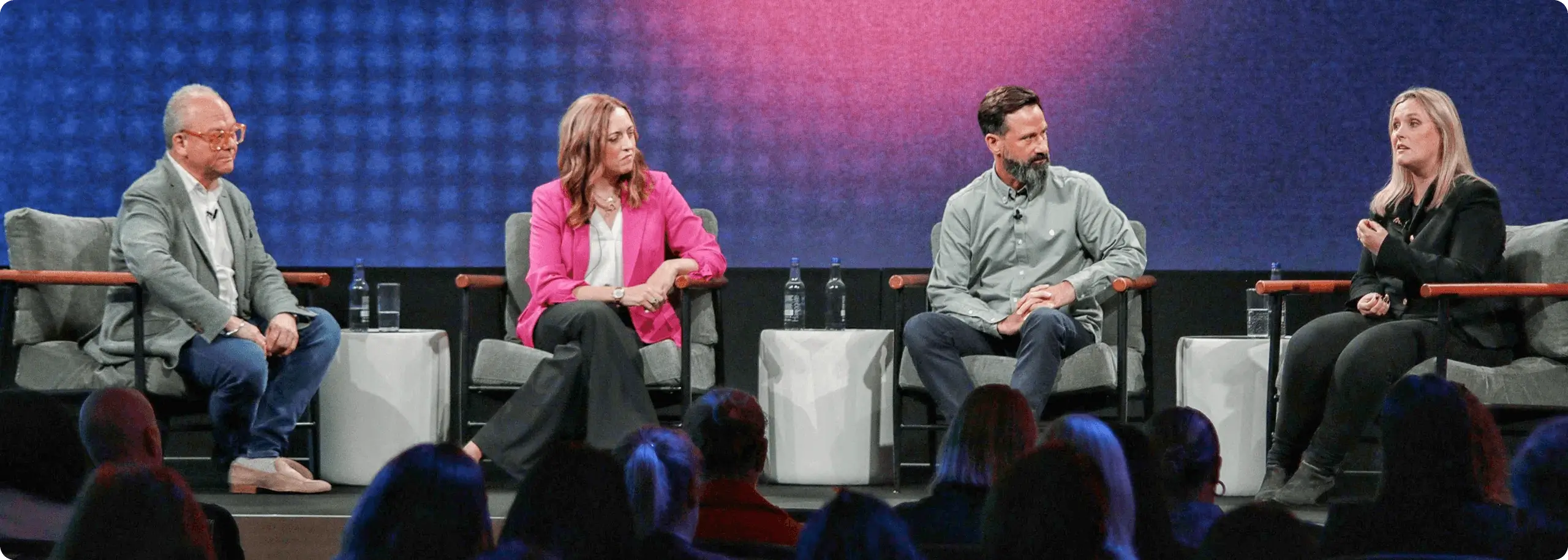 A panel of four speakers is seated on stage during a Workhuman Spotlight event. They are positioned on chairs with clear plastic water bottles on small tables beside them. The speakers are engaging with the audience, while a large, colorful backdrop sets the stage. The audience is visible in the foreground, listening attentively to the discussion.