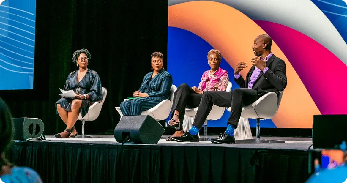 A panel of four speakers engaged in discussion at a Workhuman event. They are seated on white chairs on a stage with a colorful background. The speakers include a woman with short black hair and glasses, a woman with curly hair wearing a brightly colored top, a woman with shorter blonde hair wearing a patterned outfit, and a man with a shaved head in a suit. The audience is visible in front of them, focused on the panel conversation.