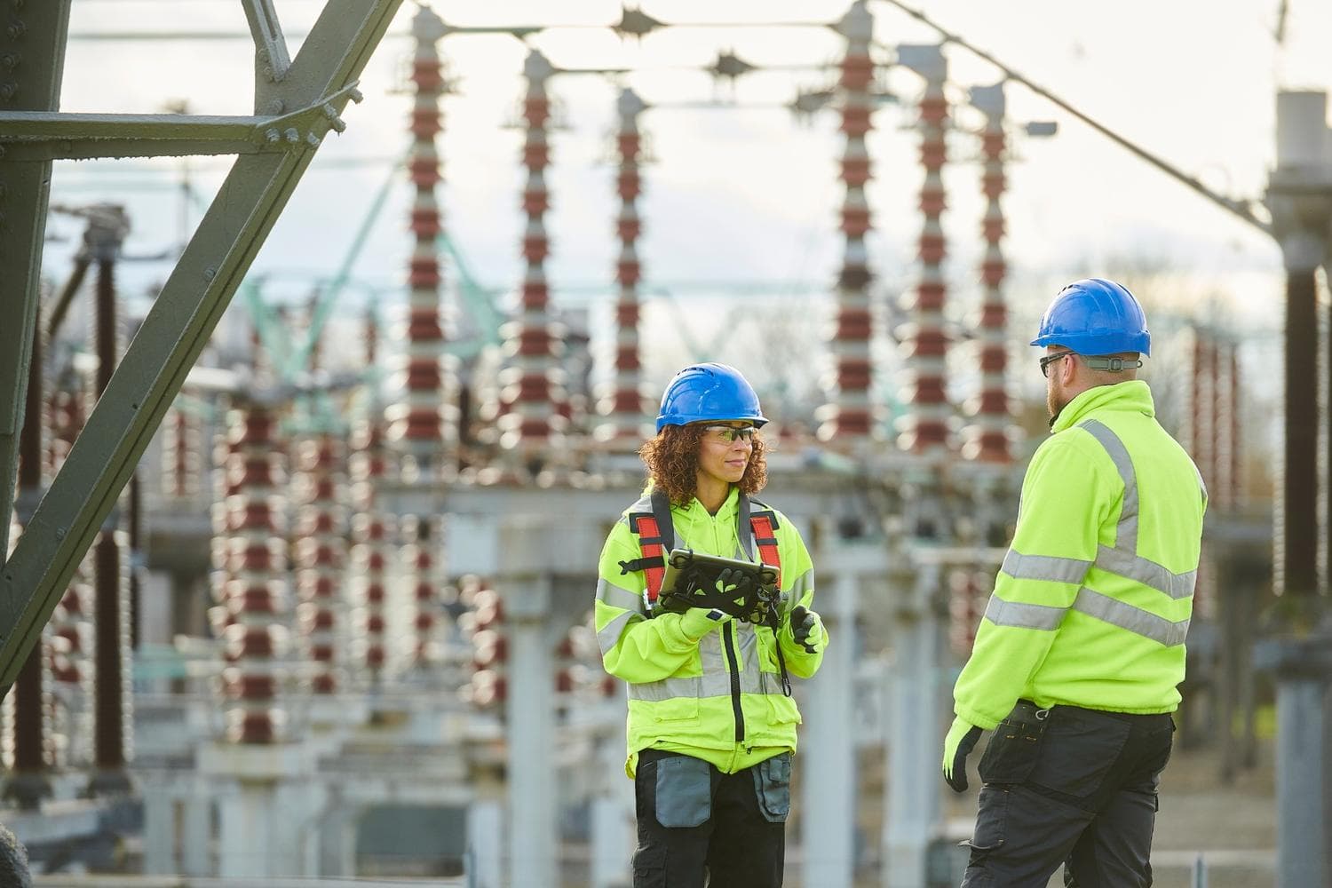 Two workers in bright yellow safety jackets and hard hats stand in a power station. One person, holding a tablet, is speaking to the other, with large electrical equipment visible in the background.