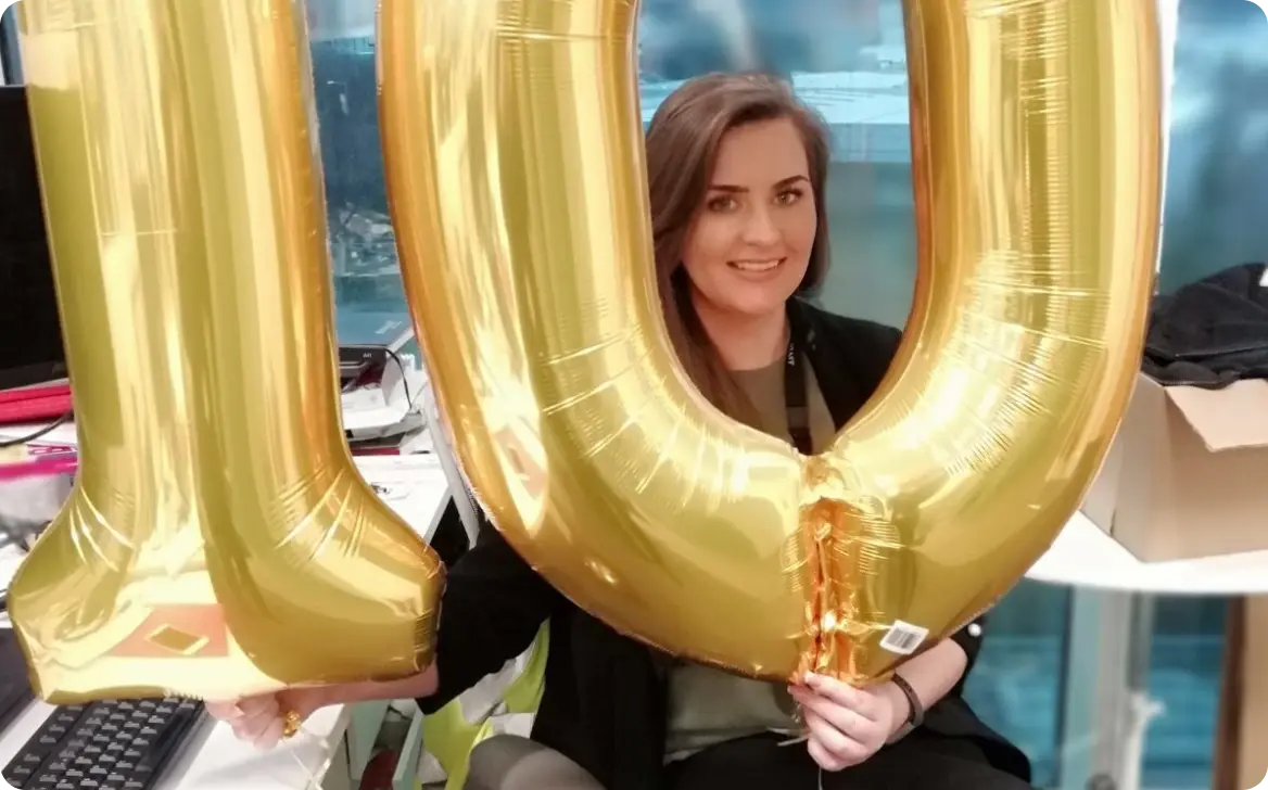 A person is smiling and holding large gold balloons that display the number "10". They are situated at a desk with various items in the background, including a computer and a box. The environment appears to be informal, suggesting a celebratory atmosphere.