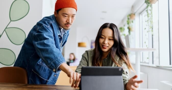 A man and a female coworker are engaged in a conversation at a table in a well-lit space, reviewing the best employee engagement platforms. The man, wearing a red beanie and a denim jacket, is pointing at a tablet in front of the woman, who is smiling and wearing a green jacket. In the background, there are additional people seated and green plants lining the windowsill.